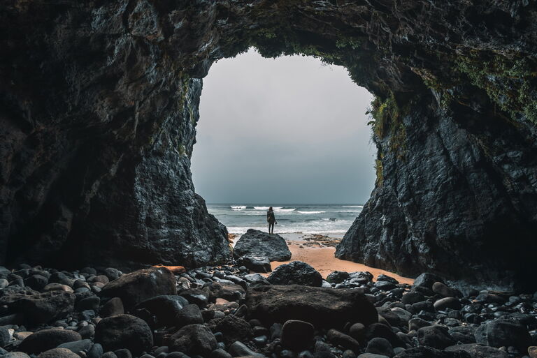 Blick aus einer Steinhöle hinaus. Der Ausgang ist riesig und vor der Höle ist das Strand und Meer zu sehen