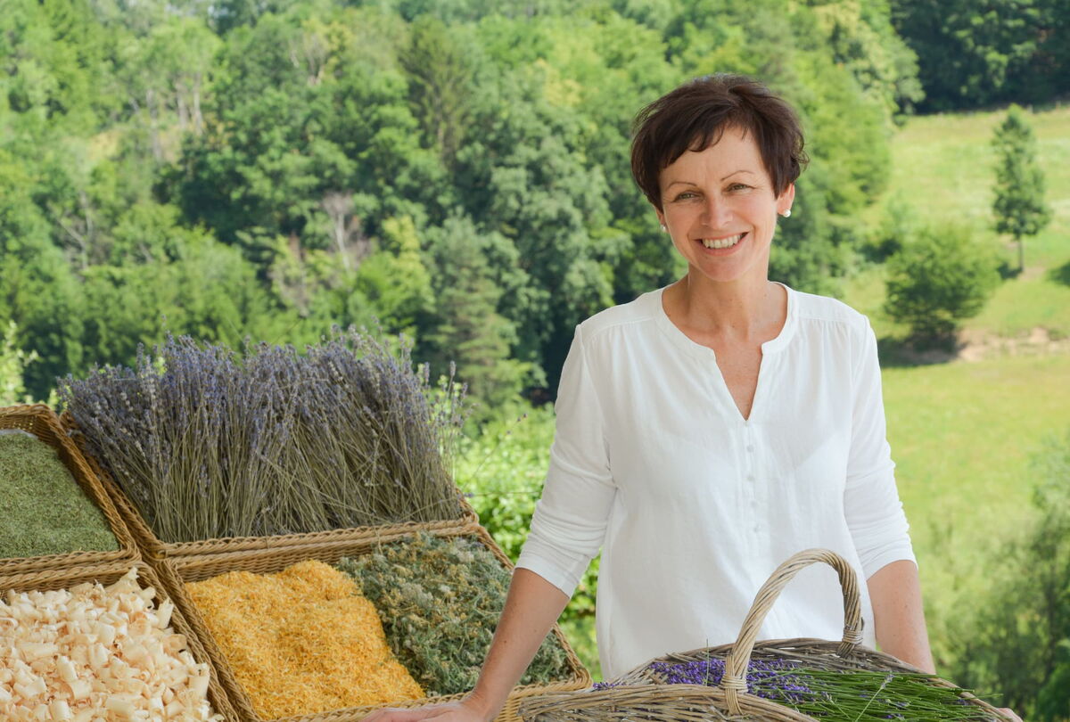 Eine Frau Präsentiert frische Kreuter in Körben, im Hintergrund ein Waldstück mit Wiese